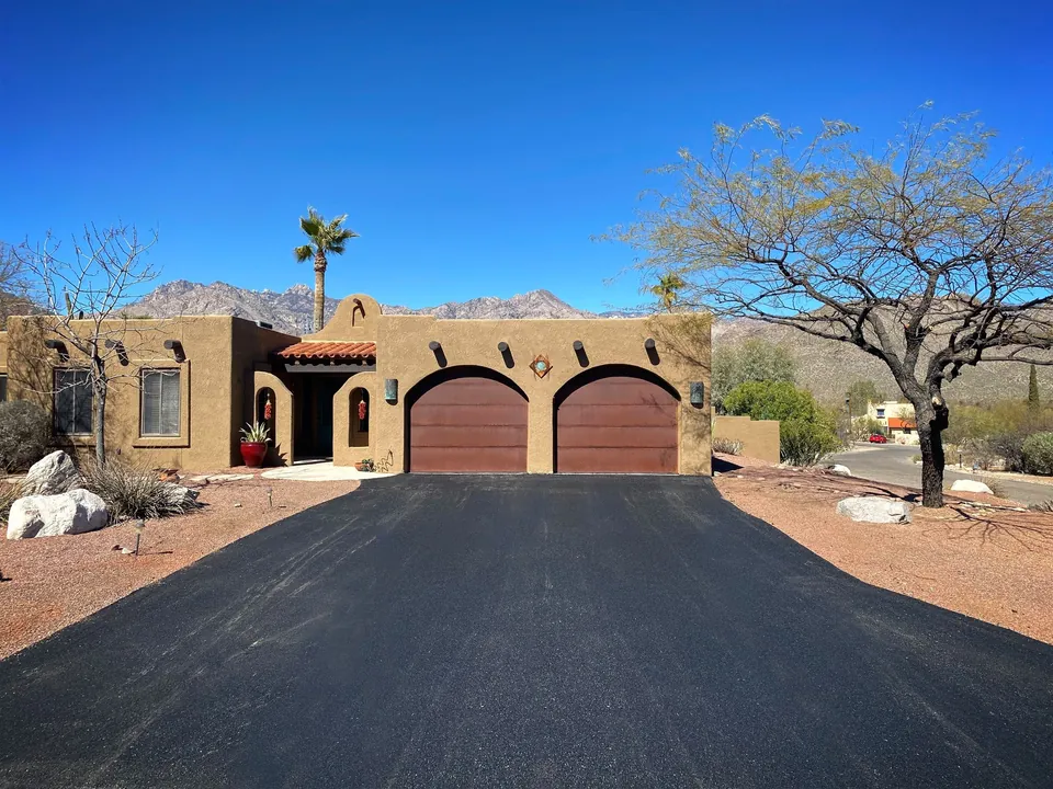 New black asphalt driveway in front of a tan adobe-style home with two arched garage doors, desert trees and rocky mountains behind
