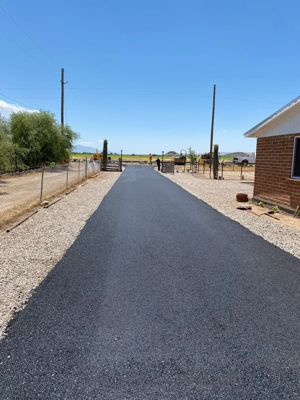 Fresh asphalt driveway between gravel shoulders and a brick home, with dry brush and desert hills beyond the fence