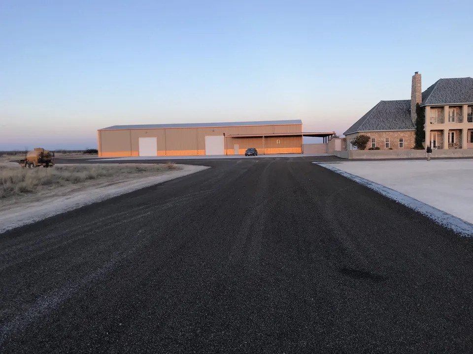 Wide new asphalt apron between a large metal shop building and a stone ranch house on open dry ground at dusk